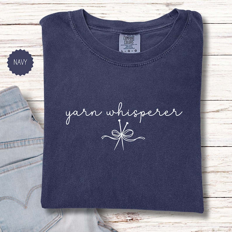 May include: Navy blue t-shirt with the words "yarn whisperer" in a cursive font and a graphic of yarn needles. The shirt is made of a soft material and is a Comfort Colors brand.