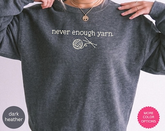 May include: A dark heather gray crewneck sweatshirt featuring the white text "never enough yarn." and a graphic of a ball of yarn with two knitting needles. The garment has long sleeves and a ribbed neckline, providing a simple and clean design.