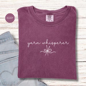 May include: Berry-colored t-shirt with the words "yarn whisperer" in white script. The shirt features a crew neck and a graphic of yarn needles and a bow. The shirt is folded on a light wood-grain surface.