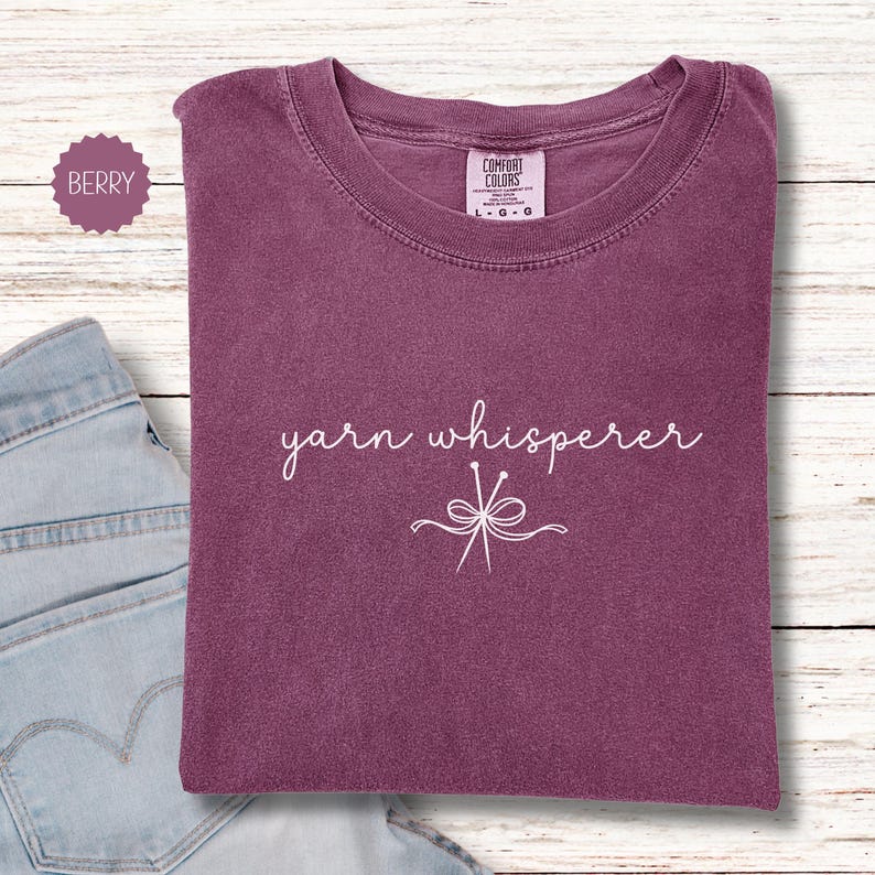 May include: Berry-colored t-shirt with the words "yarn whisperer" in white script. The shirt features a crew neck and a graphic of yarn needles and a bow. The shirt is folded on a light wood-grain surface.