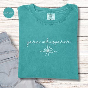 May include: Seafoam green t-shirt with the words "yarn whisperer" in white script and a graphic of knitting needles. The shirt is folded and placed on a light wood surface next to a pair of blue jeans.