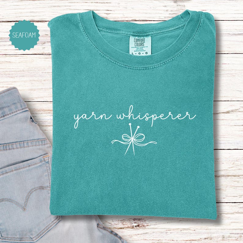 May include: Seafoam green t-shirt with the words "yarn whisperer" in white script and a graphic of knitting needles. The shirt is folded and placed on a light wood surface next to a pair of blue jeans.