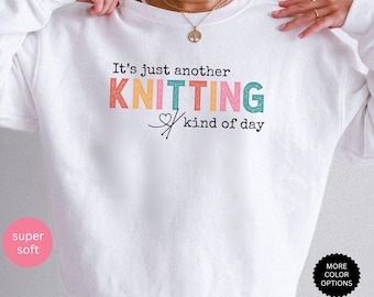 May include: A white long-sleeve sweatshirt featuring the black text It's just another KNITTING kind of day, with the word KNITTING displayed in colorful, textured block letters and a small heart icon integrated with two crossed knitting needles.