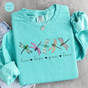 May include: A mint green crewneck sweatshirt featuring a graphic of five colorful dragonflies arranged in a horizontal row. Below the dragonflies, the black cursive text reads live, hope, grow, love, with small black hearts between each word.