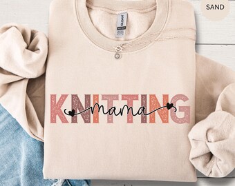 May include: A sand-colored crewneck sweatshirt featuring the word KNITTING in large, distressed, multi-colored block letters. The word mama is written in black cursive script across the center, accented with two small black hearts at each end.