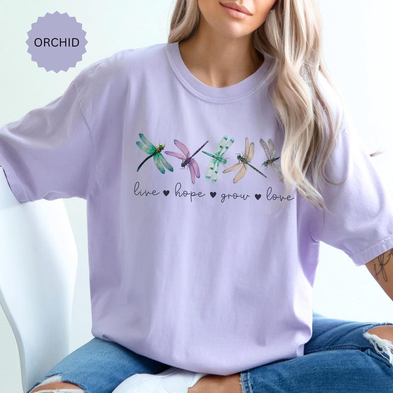 以下が含まれることがあります： 緑、紫、桃色のトンボのデザインが施された、蘭色のTシャツ。「live, hope, grow, love」の文字がトンボの下にプリントされています。柔らかい素材で作られたTシャツです。