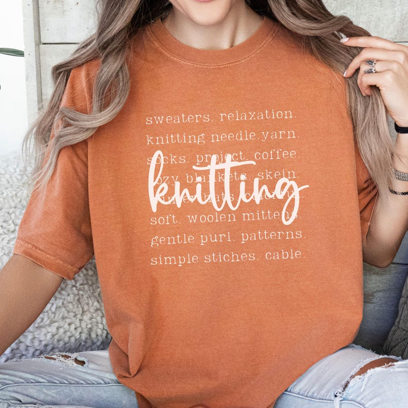 May include: Burnt orange t-shirt with the word "knitting" in a cursive font. The shirt also features a list of knitting-related words in a white font, including "sweaters," "yarn," and "patterns."