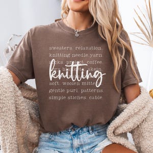 May include: A brown t-shirt with the word "knitting" in a cursive font. The shirt also features a list of knitting-related words in white, including "sweaters," "yarn," and "patterns."
