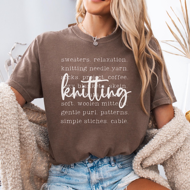 May include: A brown t-shirt with the word "knitting" in a cursive font. The shirt also features a list of knitting-related words in white, including "sweaters," "yarn," and "patterns."