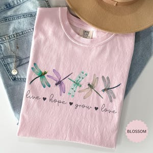以下が含まれることがあります： 5匹のカラフルなトンボのイラストが描かれた、明るいピンク色のTシャツ。「live hope grow love」の文字が下にプリントされています。ベージュの帽子と青いジーンズが背景にあります。