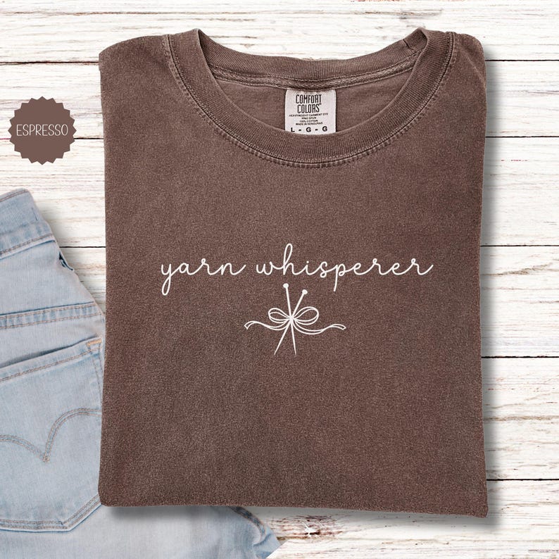 May include: Espresso-colored t-shirt with the words "yarn whisperer" in a cursive font, along with a graphic of knitting needles and yarn. The shirt is folded on a light wood surface next to a pair of blue jeans.