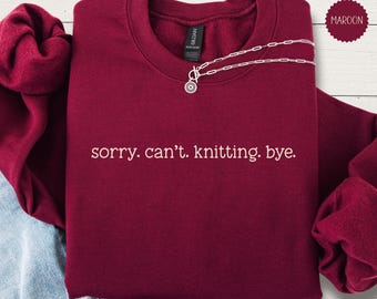 May include: A maroon crewneck sweatshirt featuring the white text "sorry. can't. knitting. bye." centered on the chest. The garment has long sleeves with ribbed cuffs and a ribbed neckline. A silver-toned chain necklace rests on the collar.
