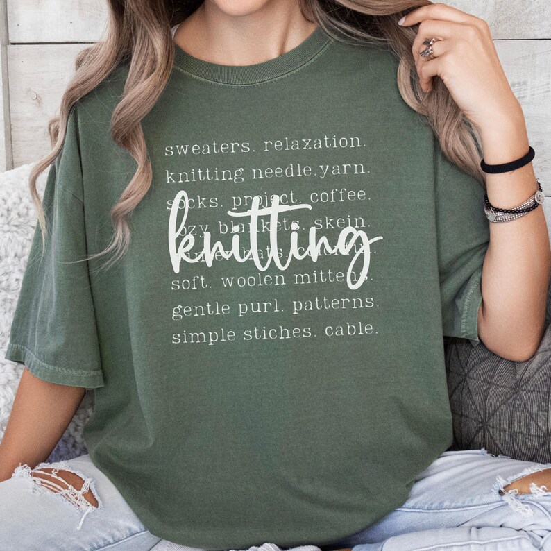 May include: Olive green t-shirt with the word "knitting" in a cursive font. The shirt features a list of knitting-related words in a white font, including "sweaters," "yarn," and "patterns."