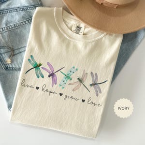 以下が含まれることがあります： 緑、紫、ピンクの色合いのトンボのデザインが施されたアイボリー色のTシャツ。「live, hope, grow, love」の文字がトンボの下にプリントされています。ベージュの帽子と青いジーンズが背景にあります。
