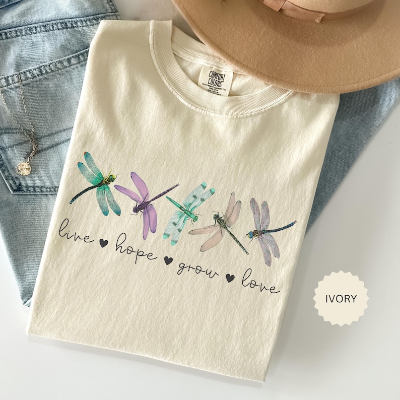 以下が含まれることがあります： 緑、紫、ピンクの色合いのトンボのデザインが施されたアイボリー色のTシャツ。「live, hope, grow, love」の文字がトンボの下にプリントされています。ベージュの帽子と青いジーンズが背景にあります。