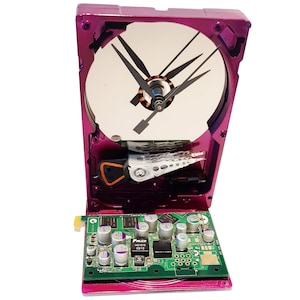 May include: A unique, handmade clock crafted from a repurposed magenta computer hard drive. The clock face is the hard drive's disc, with black clock hands. A green circuit board forms the base, creating a striking blend of technology and art.