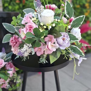 May include: A floral arrangement featuring white, pink, and lavender artificial flowers mixed with green leaves with white edges. The bouquet is displayed in a black container resting on a black metal stand with three thin legs.