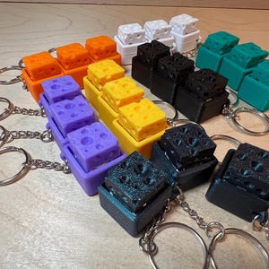 May include: A set of colorful keychain charms shaped like blocks of cheese. The charms are made of 3D printed plastic and come in a variety of colors, including orange, yellow, purple, teal, black, and green. Each charm has a small metal ring for attaching to a keychain.