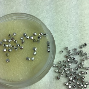May include: A small clear container filled with silver-colored beads. The beads are scattered on a white surface.