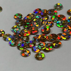 May include: A pile of iridescent gold sequins, each with a small hole in the center. The sequins are scattered on a white surface.