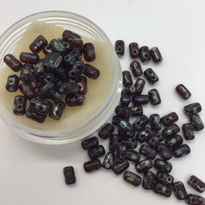Peut inclure: Un récipient de perles de verre rouge foncé et noir avec des reflets blancs et dorés. Les perles sont rectangulaires avec deux trous dans chaque perle.