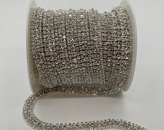 Könnte beinhalten: Eine Spule mit silberner Strass-Kettenborte. Die Kette besteht aus kleinen, quadratischen Strasssteinen, die miteinander verbunden sind. Die Kette befindet sich auf einer weißen Spule.