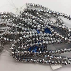 Könnte beinhalten: Eine Strähne silberfarbener, facettierter Glasperlen. Die Perlen sind klein und rund, mit einer leicht unregelmäßigen Form. Die Perlen sind auf einem durchsichtigen Faden aufgefädelt.