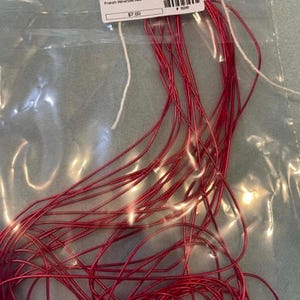 May include: A coil of thin, flexible red French wire contained within a clear plastic bag. A white rectangular label is attached to the top of the bag, featuring the text French Wire/Soft Red, a barcode, and the numerical value 7.00.