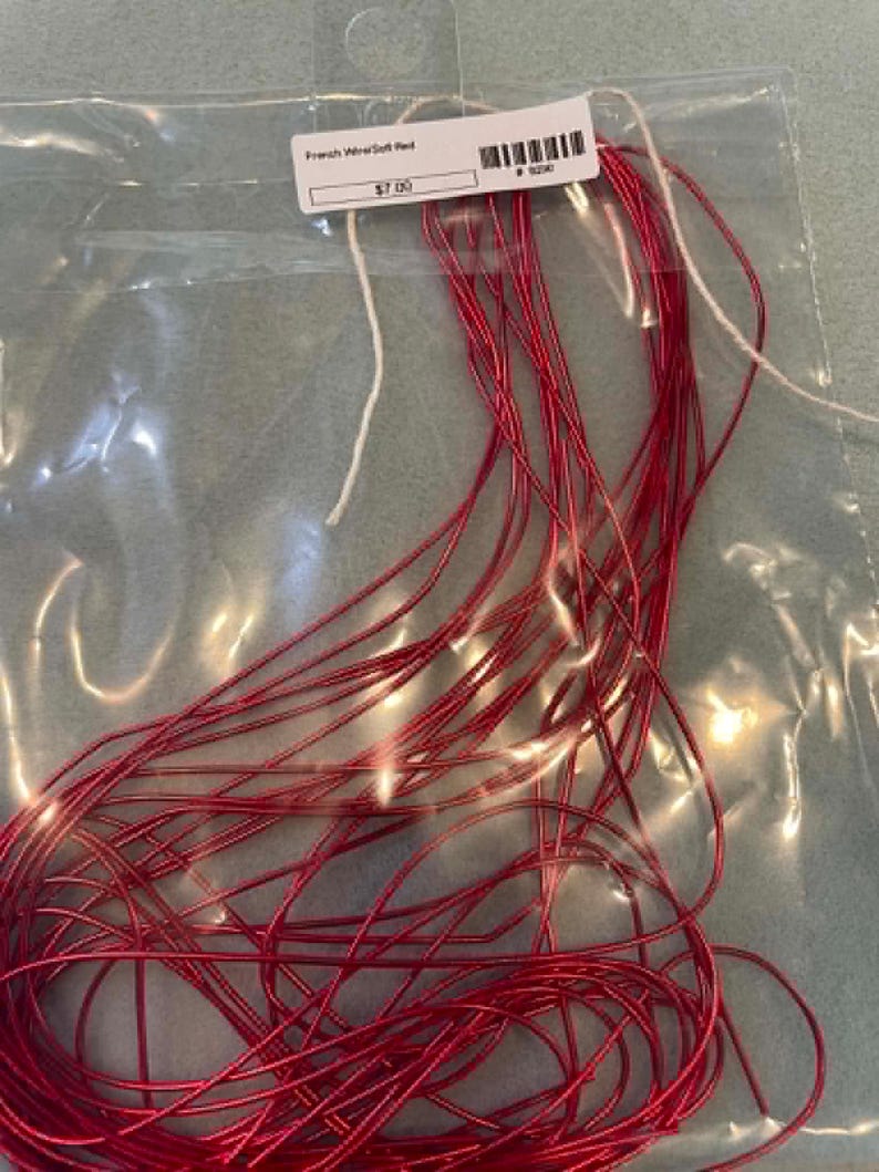 May include: A coil of thin, flexible red French wire contained within a clear plastic bag. A white rectangular label is attached to the top of the bag, featuring the text French Wire/Soft Red, a barcode, and the numerical value 7.00.