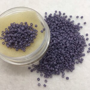 May include: A small clear container filled with purple seed beads. The beads are scattered on a white surface.