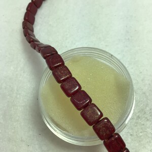 Può includere: Un filo di perline quadrate rosse con macchie dorate. Le perline sono infilate su un filo trasparente e giacciono su una superficie bianca.