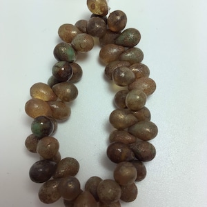 Pode incluir: Um fio de contas de vidro em forma de lágrima, de cor marrom, verde e dourado. As contas têm um acabamento fosco e estão dispostas em círculo.