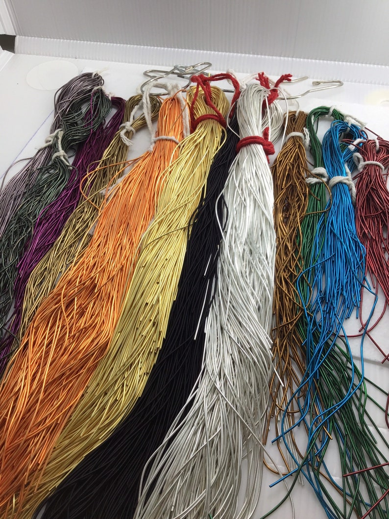 May include: A collection of thin, flexible metal wires in various colors including orange, gold, black, silver, blue, green, red, and purple. Each bundle is tied with string and hangs from a metal hook, displaying a vibrant array of metallic finishes.