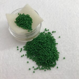 May include: A small container of green glass seed beads. The beads are scattered on a white surface.