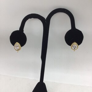 May include: A pair of gold earrings with a clear, round stone set in a halo of smaller stones. The earrings are displayed on a black velvet stand.