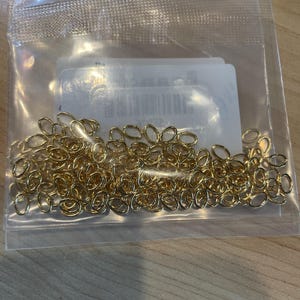 May include: A clear plastic bag filled with small, gold-colored oval chain links. The links are densely packed, creating a shiny, textured surface. The bag is partially transparent, revealing a white label with text.