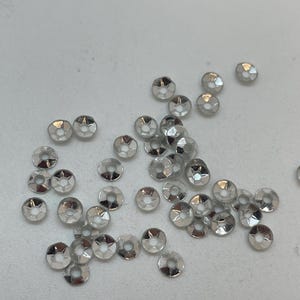 May include: A collection of small, silver-colored, flat-backed rhinestones with a central hole. These round, faceted embellishments are scattered across a light-colored surface, reflecting light and adding sparkle. Ideal for craft projects.