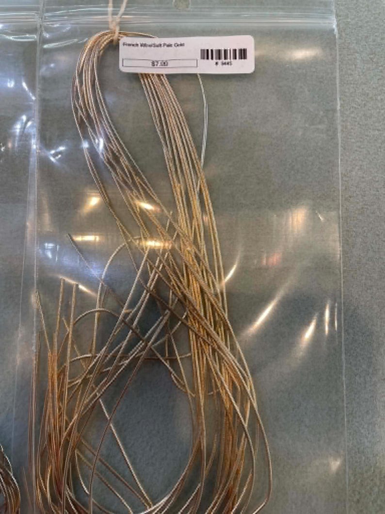 May include: A bundle of thin, metallic gold-colored French wire or gimp thread contained within a clear plastic bag. A white rectangular label is attached to the top of the bundle, featuring the text French Wire/Salt Pale Gold and a barcode.
