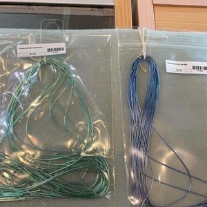 May include: Two clear plastic bags containing thin, metallic-looking cord or thread. The left bag holds a mint green cord labeled French Soft Nice Mint Green, and the right bag holds a blue cord labeled French Woven Light Blue.