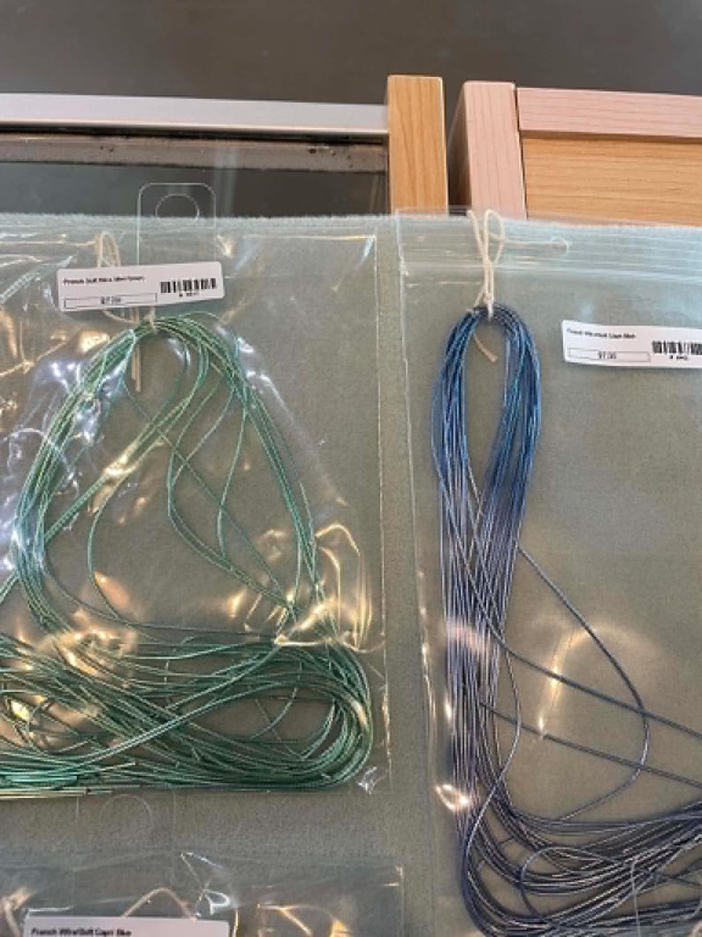 May include: Two clear plastic bags containing thin, metallic-looking cord or thread. The left bag holds a mint green cord labeled French Soft Nice Mint Green, and the right bag holds a blue cord labeled French Woven Light Blue.