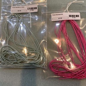 May include: Two clear plastic bags containing coiled French wire. The left bag holds light green wire with a label reading French Gimp Wire Lt. Green. The right bag holds bright pink wire with a label reading French Wire Soft Pink.