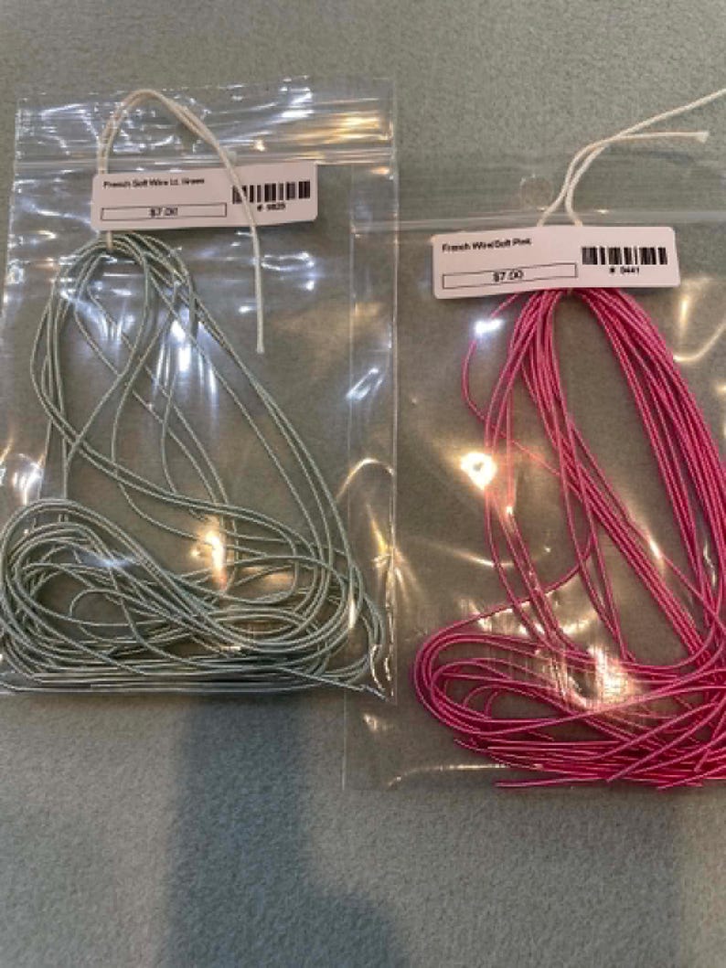May include: Two clear plastic bags containing coiled French wire. The left bag holds light green wire with a label reading French Gimp Wire Lt. Green. The right bag holds bright pink wire with a label reading French Wire Soft Pink.