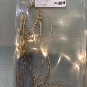 May include: A bundle of thin, flexible gold-colored French wire coiled inside a clear plastic bag. A white rectangular label is attached to the top of the bag, featuring the text French Wire/Soft Old Gold and a barcode.