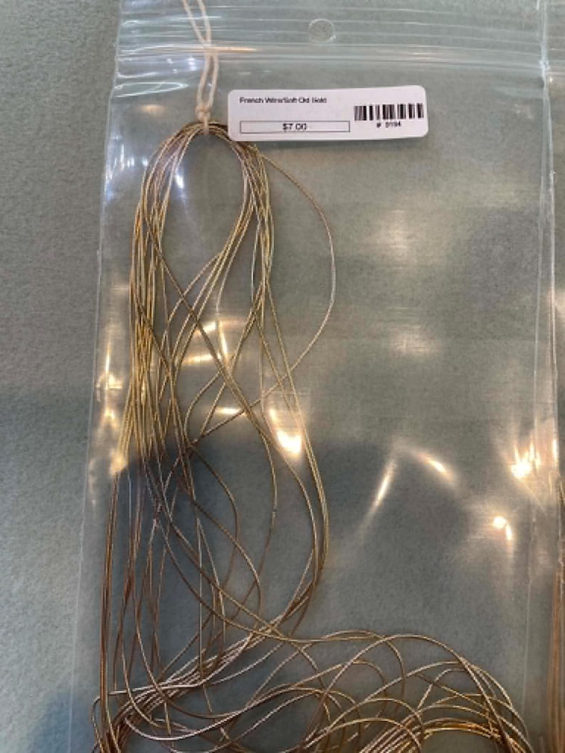 May include: A bundle of thin, flexible gold-colored French wire coiled inside a clear plastic bag. A white rectangular label is attached to the top of the bag, featuring the text French Wire/Soft Old Gold and a barcode.
