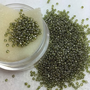 May include: A close-up of a container filled with small, round, green glass beads. The beads are scattered on a white surface.