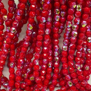 May include: Multiple strands of vibrant red faceted glass beads arranged vertically. Each bead features a polished, multi-sided surface with an iridescent finish that reflects light, creating a shimmering effect across the collection of round beads.