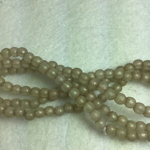 May include: A strand of light brown glass beads with a shimmery finish. The beads are round and have a slightly irregular shape.