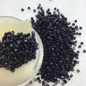 Peut inclure: Un pot en verre transparent rempli de perles noires, à côté d'un tas de perles noires en vrac. Les perles sont petites, cylindriques et ont une finition brillante. Le pot est posé sur une surface blanche.