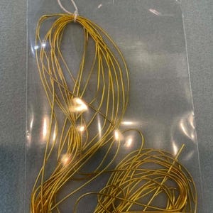 May include: A coil of thin, metallic gold-colored wire packaged in a clear plastic bag. The wire is looped and secured with a white string at the top. A white rectangular label is attached to the top of the bag with the text French Wire/Soft Saffron.