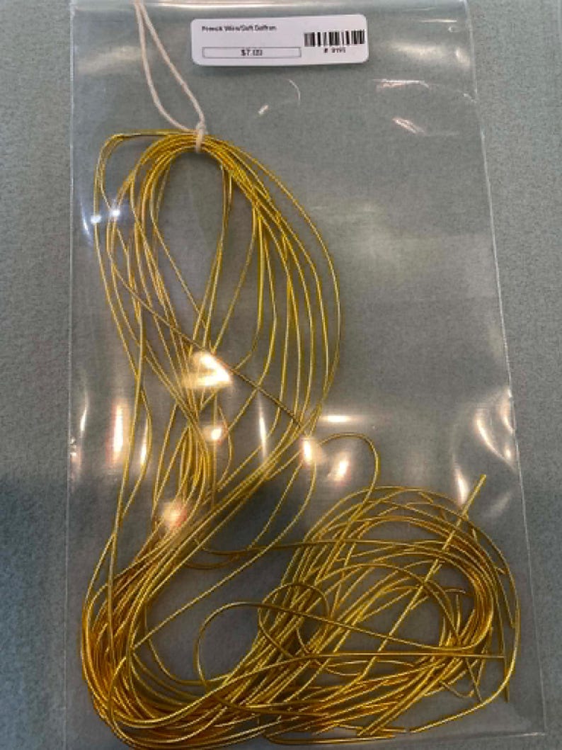 May include: A coil of thin, metallic gold-colored wire packaged in a clear plastic bag. The wire is looped and secured with a white string at the top. A white rectangular label is attached to the top of the bag with the text French Wire/Soft Saffron.